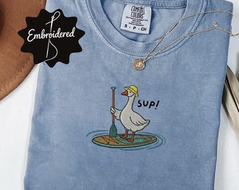 Könnte beinhalten: Ein blaues T-Shirt mit Rundhalsausschnitt und einer Stickerei einer weißen Gans mit gelbem Hut, die auf einem Stand-Up-Paddleboard steht und ein Paddel hält, mit dem in Schwarz gestickten Text "sup!" neben der Gans. Das Shirt hat ein S-Größenetikett.