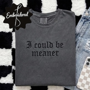 May include: A charcoal gray t-shirt with the embroidered text "I could be meaner" is paired with light wash distressed jeans and a phone case with a beige and white checkerboard pattern. The items are arranged on a black and white checkered surface.