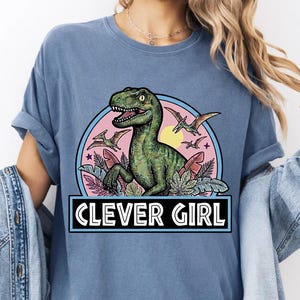 Può includere: Una t-shirt blu a maniche corte con la grafica di un dinosauro verde circondato da elementi circolari rosa e blu, foglie tropicali e pterodattili in volo. Il testo CLEVER GIRL è stampato in bianco all'interno di un rettangolo nero.