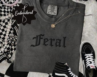 Könnte beinhalten: Ein anthrazitfarbenes Kurzarm-T-Shirt mit dem Wort Feral, das in einer schwarzen gotischen Schrift auf der Brust gestickt ist. Das T-Shirt hat einen Rundhalsausschnitt und ein Comfort Colors Markenetikett am Innenkragen für Größe S.