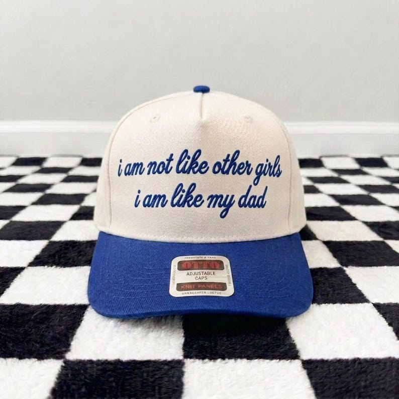 K&ouml;nnte beinhalten: Eine cremefarbene Baseballkappe mit blauem Schirm und blauem gesticktem Text auf der Vorderseite, der lautet: "i am not like other girls i am like my dad." Die Kappe hat einen kleinen blauen Knopf oben und einen rechteckigen Aufkleber auf dem Schirm.