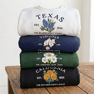 May include: A stack of four crewneck sweatshirts featuring embroidered state names, establishment years, and floral designs. The white sweatshirt shows Texas bluebonnets, navy shows Virginia dogwood, green shows Cherokee roses, and black shows California poppies.