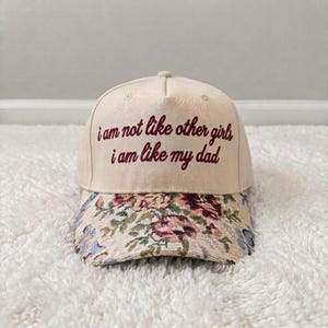 K&ouml;nnte beinhalten: Eine beige Baseballkappe mit dem gestickten Text "i am not like other girls i am like my dad" in dunkelroter Schreibschrift. Der Schirm der Kappe ist mit einem gewebten Blumenmuster in Rosa-, Rot-, Gr&uuml;n- und Blaut&ouml;nen verziert.
