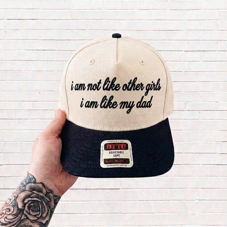 K&ouml;nnte beinhalten: Eine zweifarbige Baseballkappe mit cremefarbener Krone und schwarzem Schirm. Auf der Vorderseite befindet sich ein schwarzer Sticktext mit der Aufschrift: i am not like other girls i am like my dad. Ein rechteckiger Aufkleber auf dem Schirm zeigt den Markennamen OTTO.