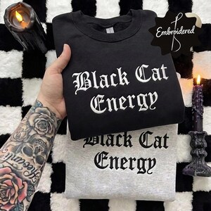 Puede incluir: Se muestran dos sudaderas, una negra y otra gris. La sudadera negra tiene bordadas las palabras "Black Cat Energy" en letras góticas blancas. La sudadera gris también tiene el mismo texto. El fondo es un patrón de cuadros blancos y negros.