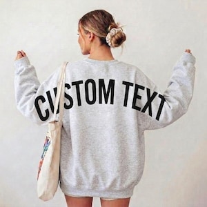 May include: A light gray crewneck sweatshirt featuring the words CUSTOM TEXT printed in large black capital letters across the back. The sweatshirt has long sleeves and a relaxed fit, paired with a beige canvas tote bag hanging from one shoulder.