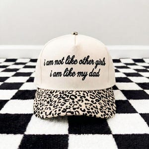 K&ouml;nnte beinhalten: Eine cremefarbene Baseballkappe mit einem Leopardenmuster-Schirm und dem in Schwarz gestickten Text "i am not like other girls i am like my dad" auf der Vorderseite. Die Kappe hat eine strukturierte Krone und einen Knopf an der Oberseite.