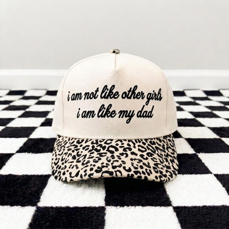 K&ouml;nnte beinhalten: Eine cremefarbene Baseballkappe mit einem Leopardenmuster-Schirm und dem in Schwarz gestickten Text "i am not like other girls i am like my dad" auf der Vorderseite. Die Kappe hat eine strukturierte Krone und einen Knopf an der Oberseite.
