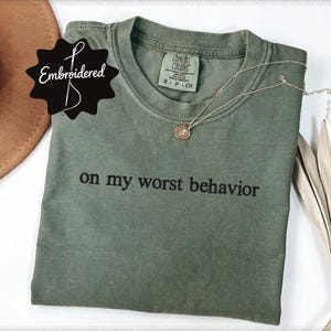 May include: An olive green short-sleeve t-shirt with the text "on my worst behavior" embroidered in black thread across the chest. The shirt features a crew neckline and a Comfort Colors brand label inside the collar, size small.