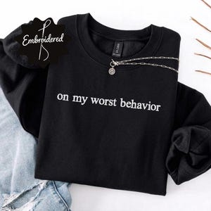 May include: A black crewneck sweatshirt featuring the white embroidered text "on my worst behavior" across the chest. The garment has long sleeves and a ribbed neckline, with a silver-toned chain necklace resting on the collar area.