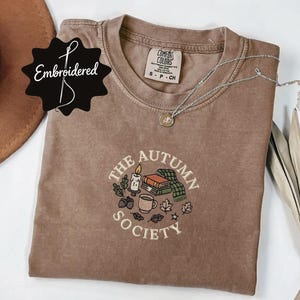 Könnte beinhalten: Ein braunes kurzärmeliges T-Shirt mit Rundhalsausschnitt und einer gestickten Grafik auf der Brust. Das Motiv zeigt eine Kerze, einen Bücherstapel, eine Tasse und Blätter mit dem Text "THE AUTUMN SOCIETY" in kreisförmiger Anordnung.