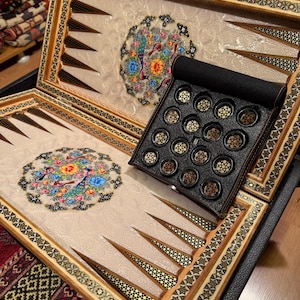 May include: Backgammon board with a decorative floral design. The board has a wooden frame with inlaid patterns and a black leather case. The game pieces are black and gold.