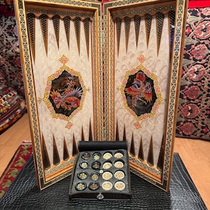 May include: A wooden backgammon board featuring intricate inlaid geometric borders and two central panels depicting colorful phoenix illustrations. The set includes a black leather-textured carrying case and a tray containing dark and light game pieces.