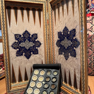 Könnte beinhalten: Backgammon-Set mit Holzrahmen, cremefarbener Spielfläche und blauen und goldenen floralen Designs. Ein schwarzes Lederetui enthält Spielsteine und Würfel. Das Spielbrett ist offen und spielbereit. Ein klassisches Brettspiel.