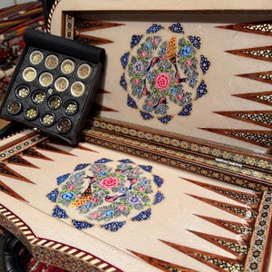 Könnte beinhalten: Ein detailliertes Backgammon-Set mit cremefarbener Spielfläche, verziert mit einem zentralen Blumendesign mit Vögeln in Rosa, Blau und Grün. Das Set enthält eine schwarze Box mit goldenen und schwarzen Spielsteinen und einen dekorativen Holzrahmen.