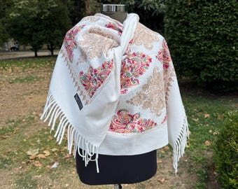 May include: A white shawl with a paisley pattern in shades of tan, red, and green. The shawl has a fringed edge and is displayed on a mannequin. The background features green foliage.