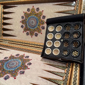 Könnte beinhalten: Backgammon-Spielbrett mit cremefarbener Spielfläche, mit einem zentralen Blumendesign in Blau, Gold und Rot. Der Holzrahmen ist mit dekorativen Mustern eingelegt. Ein schwarzes Lederetui enthält die Spielsteine, die gold und schwarz sind.