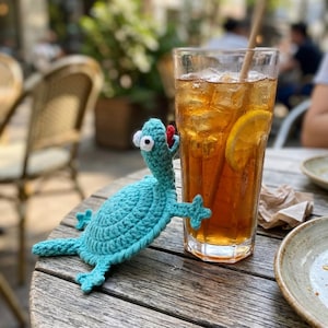 May include: A turquoise crocheted turtle toy with large white and black googly eyes and a small red tongue, positioned next to a tall glass filled with iced tea, ice cubes, a lemon slice, and a straw on a wooden table surface.