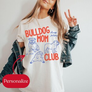 May include: Cream-colored t-shirt with the words "BULLDOG MOM CLUB" in red and blue. The shirt features three line drawings of bulldogs and the phrase "always judging." The text "est. 2025" is also included.