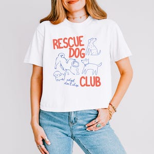 May include: A white short-sleeved t-shirt featuring the red text "RESCUE DOG CLUB" and the blue text "adopt don't shop". The graphic includes blue line-art illustrations of four different dogs and a bone, centered on the front of the shirt.