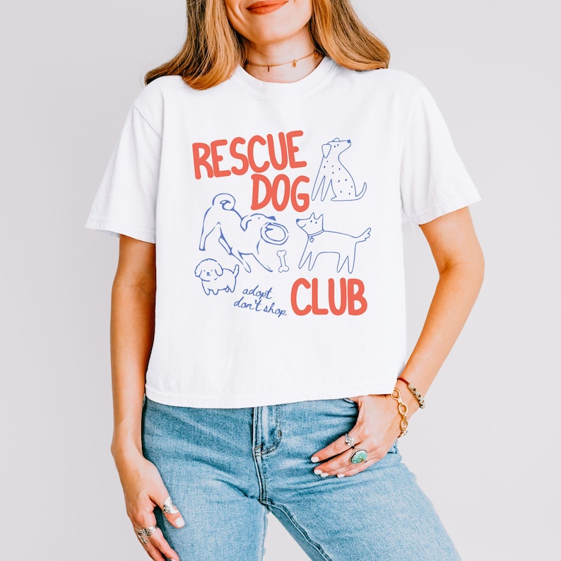 May include: A white short-sleeved t-shirt featuring the red text "RESCUE DOG CLUB" and the blue text "adopt don't shop". The graphic includes blue line-art illustrations of four different dogs and a bone, centered on the front of the shirt.