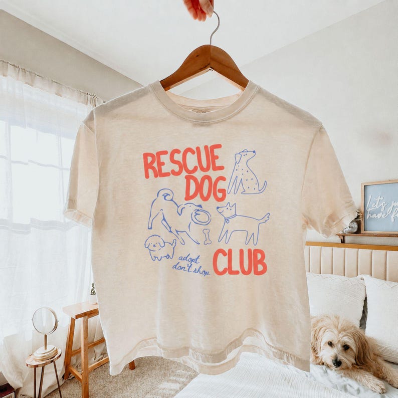 May include: A cream-colored short-sleeve t-shirt featuring the red text "RESCUE DOG CLUB" and blue line-art illustrations of various dogs. The graphic includes the phrase "adopt don't shop" in blue script and a small bone icon.