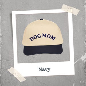 May include: A baseball cap with a cream-colored crown and a navy blue brim. The words "DOG MOM" are embroidered in navy blue across the front. The cap has a curved brim and a small button on top. The word "Navy" is written below the cap.