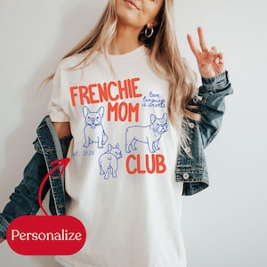 May include: Cream-colored t-shirt with the words "FRENCHIE MOM CLUB" in red, along with blue illustrations of French Bulldogs. The text "love language is snorts" is also present. The shirt has the text "est. 2025" on it.