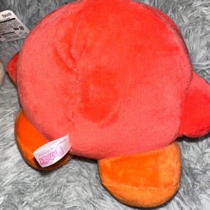 May include: A plush toy in the shape of a character, primarily red with orange feet. The toy has a soft, fuzzy texture and a tag attached. The background is a gray, fuzzy surface.