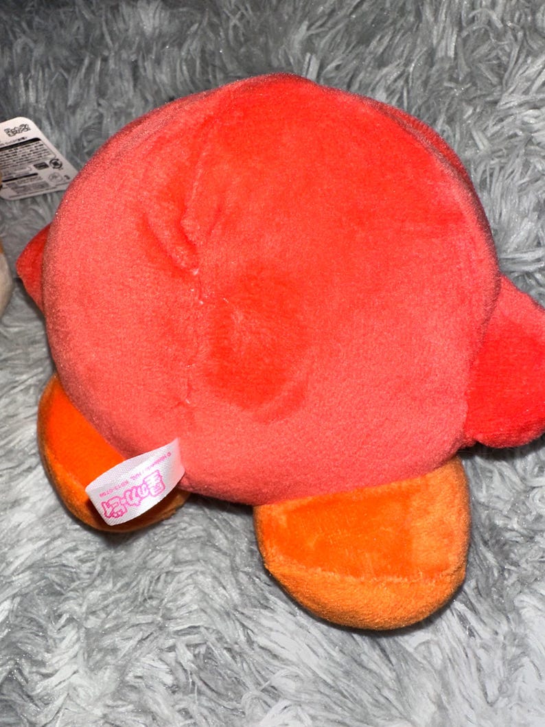 May include: A plush toy in the shape of a character, primarily red with orange feet. The toy has a soft, fuzzy texture and a tag attached. The background is a gray, fuzzy surface.