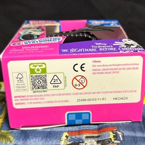 Puede incluir: Una caja rosa y azul con texto de The Nightmare Before Christmas de Tim Burton. La caja incluye advertencias, informaci&oacute;n de reciclaje y el logotipo de Disney. Contiene una mochila de juguete y accesorios sorpresa.