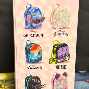 Puede incluir: Una caja rosa con mini mochilas tem&aacute;ticas de Disney. Los dise&ntilde;os incluyen Lilo & Stitch, Disney Princess, Moana, Villains, The Nightmare Before Christmas y Mickey Mouse. El texto "Collect them all!" se muestra en la parte superior.