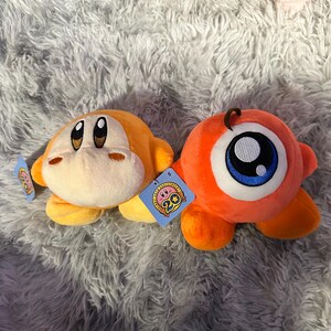 May include: Two plush toys: an orange character with a tan face and a large eye character. The orange character has a tag with a pink and blue design. The eye character has a large blue and white eye. Both are on a gray, fluffy surface.