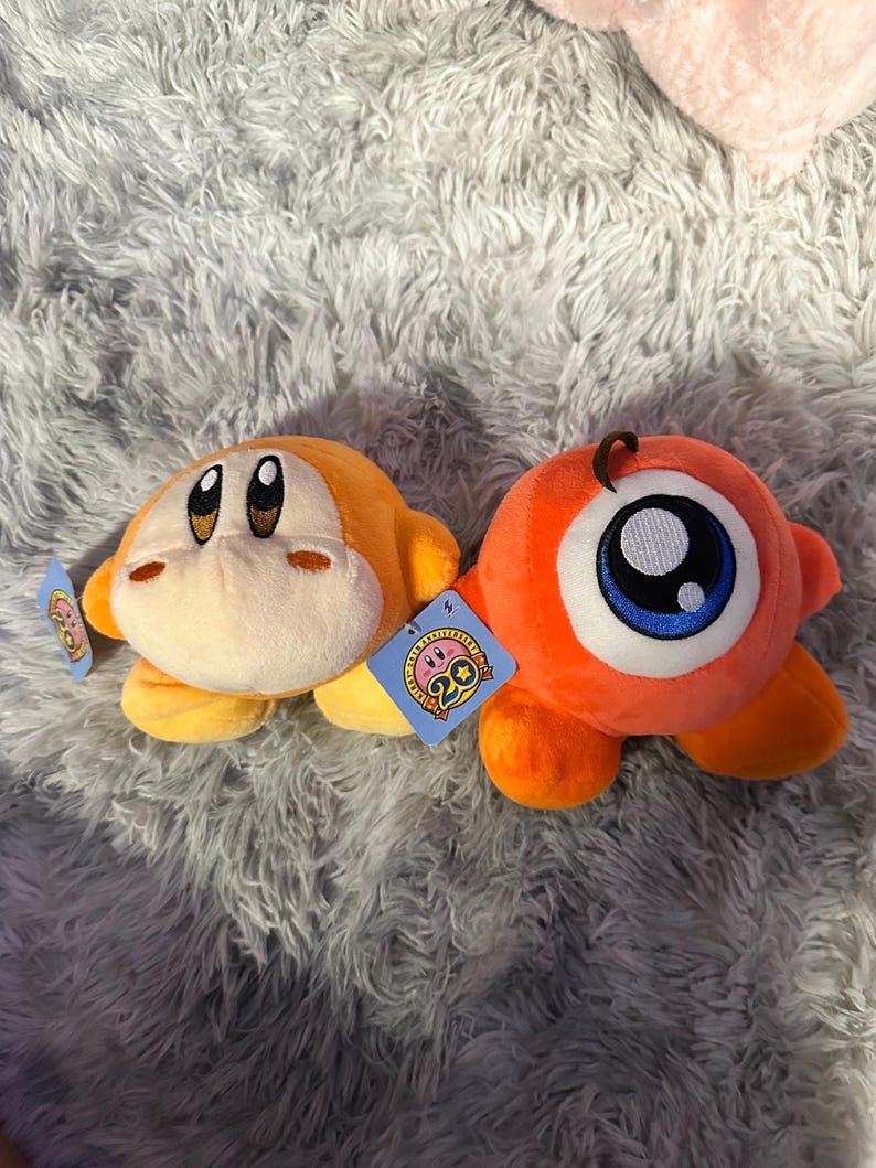 May include: Two plush toys: an orange character with a tan face and a large eye character. The orange character has a tag with a pink and blue design. The eye character has a large blue and white eye. Both are on a gray, fluffy surface.