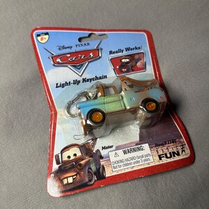 May include: A Disney Pixar Cars Mater light-up keychain in original blister packaging. The packaging features the Cars logo, the text Light-Up Keychain, and a warning label. The keychain is a miniature blue and brown tow truck with orange wheels.