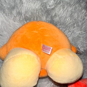 May include: An orange plush toy with two light yellow rounded shapes and a small tag. The toy is on a gray, fluffy surface. The plush toy is a vibrant orange color.