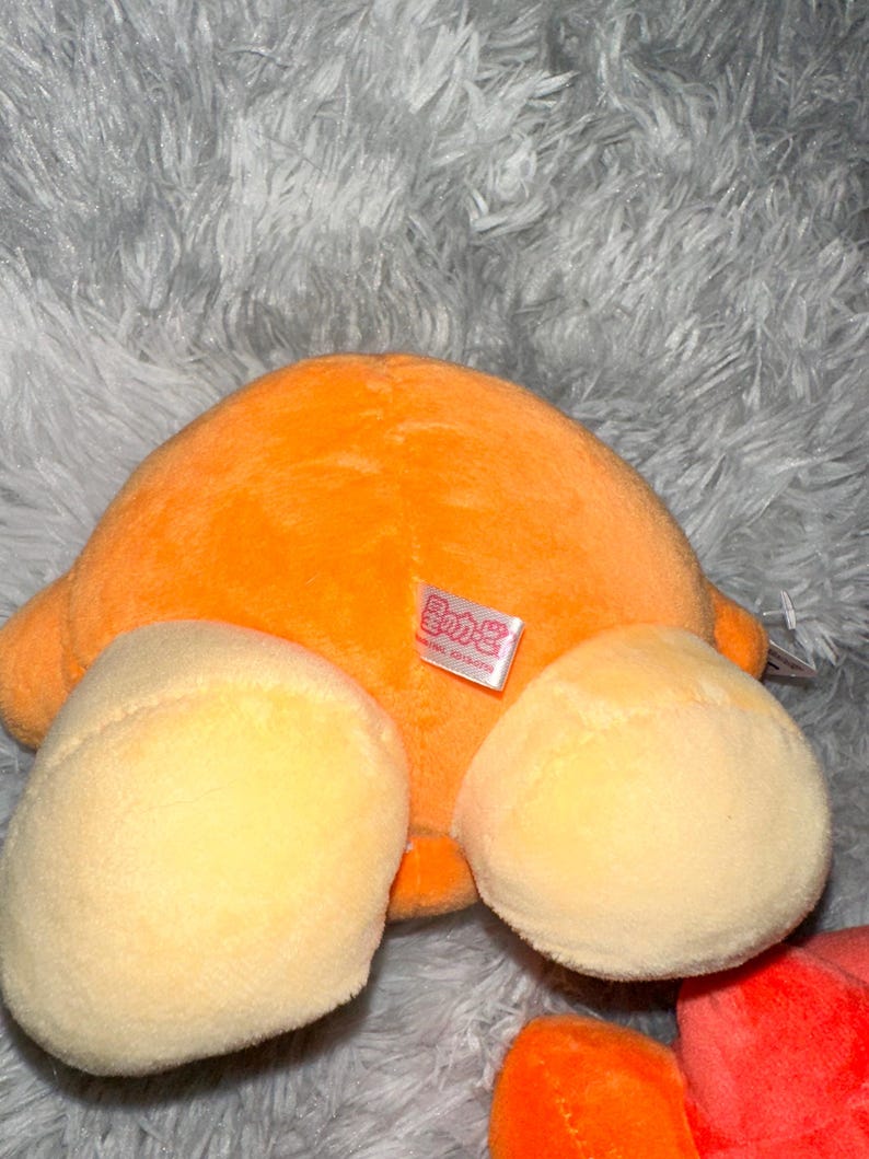 May include: An orange plush toy with two light yellow rounded shapes and a small tag. The toy is on a gray, fluffy surface. The plush toy is a vibrant orange color.