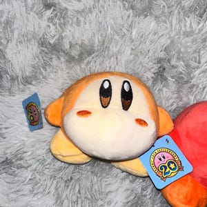 May include: A plush toy of a character with a round body, orange skin, and large black eyes. The toy has a tag that reads "20th Anniversary". The plush is on a fluffy, gray surface.