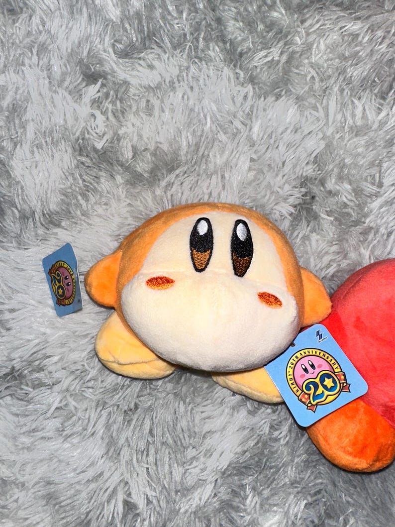 May include: A plush toy of a character with a round body, orange skin, and large black eyes. The toy has a tag that reads "20th Anniversary". The plush is on a fluffy, gray surface.