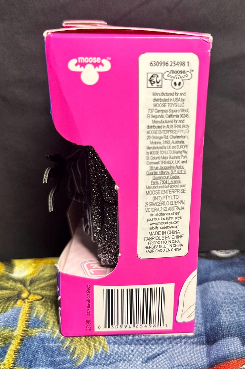 Puede incluir: Una caja de producto rosa y negra con el logotipo de Moose Toys. La caja tiene un c&oacute;digo de barras e informaci&oacute;n del producto impresa. El producto dentro de la caja es parcialmente visible.