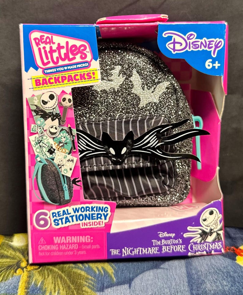 Puede incluir: Una mochila Real Littles Disney The Nightmare Before Christmas en una caja. La mochila es negra con purpurina plateada y un dise&ntilde;o de murci&eacute;lago Jack Skellington. La caja tiene el logotipo de Disney y el texto "Real Working Stationery Inside!"