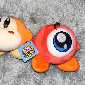 May include: Two plush toys, one orange and one red, rest on a gray, fuzzy surface. The orange toy has a round face and small arms. The red toy has a large eye with blue and black details. A tag with a pink character and the number 20 is attached.
