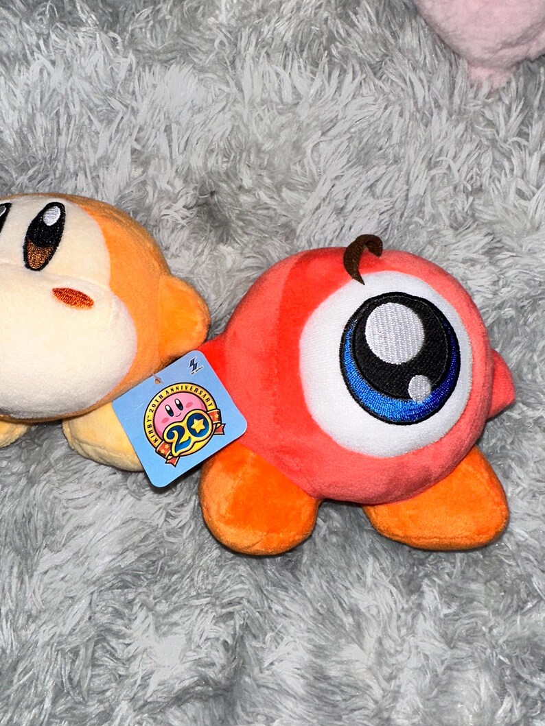 May include: Two plush toys, one orange and one red, rest on a gray, fuzzy surface. The orange toy has a round face and small arms. The red toy has a large eye with blue and black details. A tag with a pink character and the number 20 is attached.