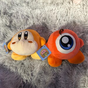 May include: Two plush toys, one orange and one yellow, rest on a fluffy gray surface. The orange toy has a large eye, while the yellow one has a smiling face. Both have tags attached, and the toys are part of a collection.