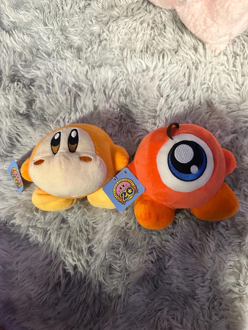 May include: Two plush toys, one orange and one yellow, rest on a fluffy gray surface. The orange toy has a large eye, while the yellow one has a smiling face. Both have tags attached, and the toys are part of a collection.