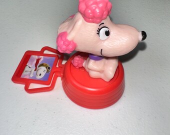May include: A pink poodle figurine with a red base and a small red frame featuring a cartoon image. The poodle has a pink pompadour, a black nose, and a purple collar. The figurine is a collectible item.