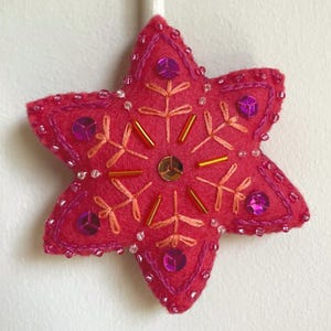 May include: A handmade, star-shaped Christmas ornament in shades of pink. The felt ornament is embellished with sequins, beads, and embroidery in complementary colors. A white ribbon is attached for hanging.