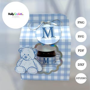 Peut inclure: Une boîte cadeau à carreaux bleus et blancs avec une découpe d'ours en peluche et un pot de miel. La boîte a une étiquette circulaire avec la lettre "M". Le pot porte également la lettre "M". La boîte est décorée d'une ficelle argentée.