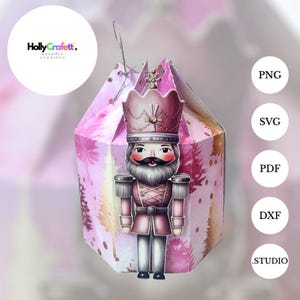 Puede incluir: Una caja de regalo hexagonal rosa y dorada con un diseño de soldado cascanueces. El cascanueces tiene un uniforme rosa, una barba blanca y una corona. La caja tiene un efecto acuarela con detalles dorados y el texto "HollyCrafett" en un círculo.
