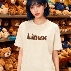 Peut inclure: Un t-shirt crème avec le mot "Linux" en lettres marron, avec des visages d'ours en peluche remplaçant les lettres "i". L'arrière-plan présente des étagères remplies d'ours en peluche.